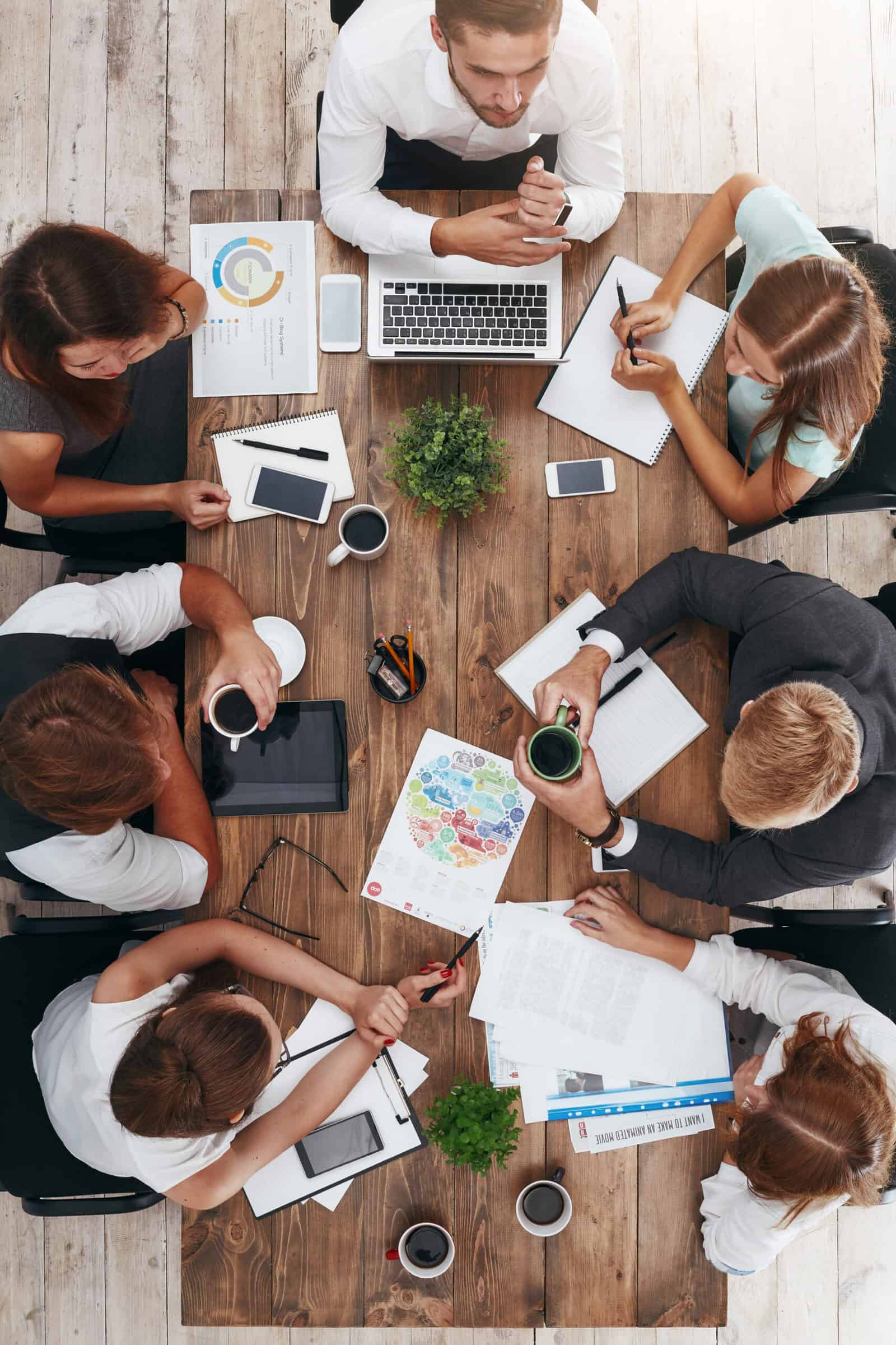 Top view of meeting for the business team, brainstorming on the new project. Laptop, potted plants, diary, coffee cups, glasses, charts and other supplies on brown wooden desk. Colleagues discussing ideas while drinking coffee in the office.